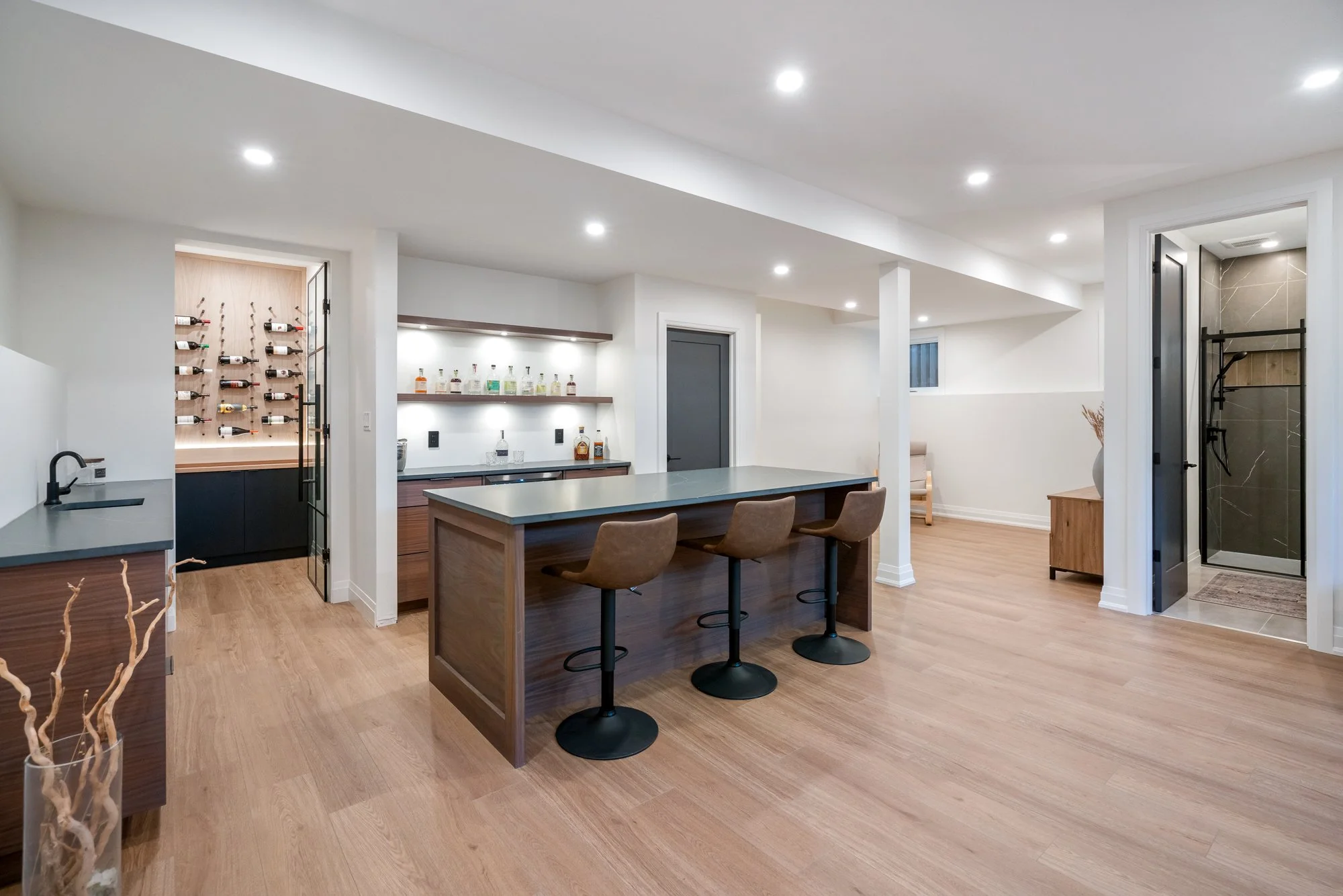 Modern recreational room with a wooden floor, a kitchen island with three brown bar stools, a bar area with a variety of liquor bottles on illuminated shelves, and a wine storage nook with bottles stored on a wall-mounted rack.