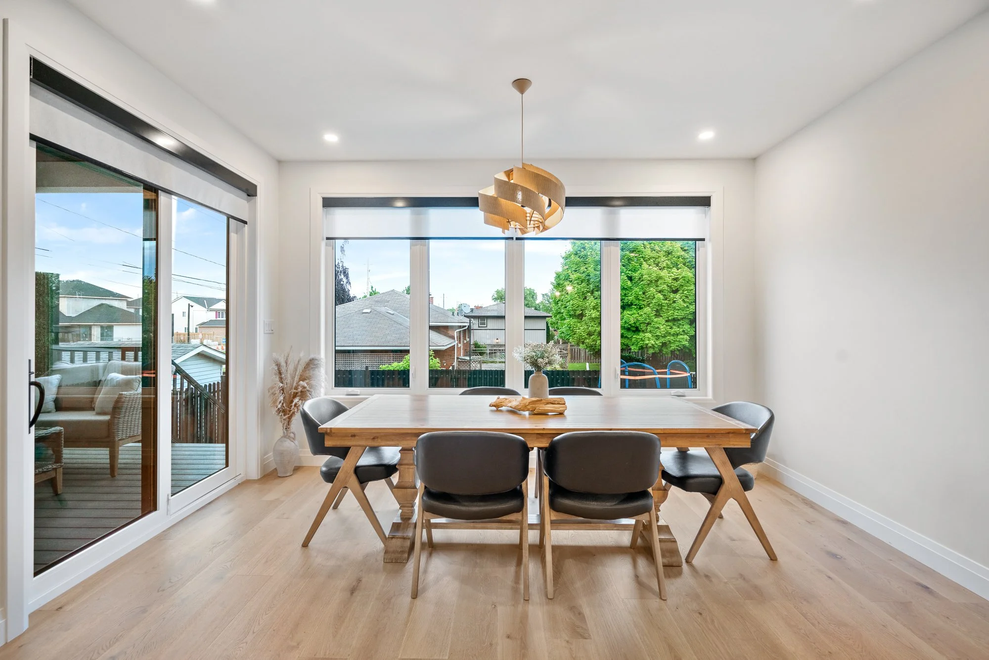 Empty dining room with wooden table, six black chairs, white walls, large windows showing outdoor greenery, sliding glass door to patio, modern light fixture hanging from ceiling, light wood flooring.