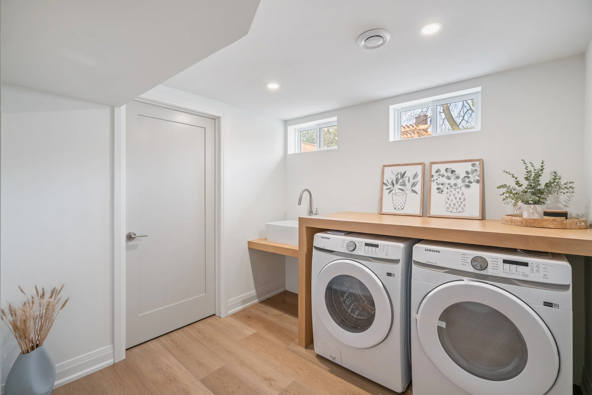 Laundry room with white washing machine and dryer under a wooden counter, a white sink, small windows, some framed art, and plants.