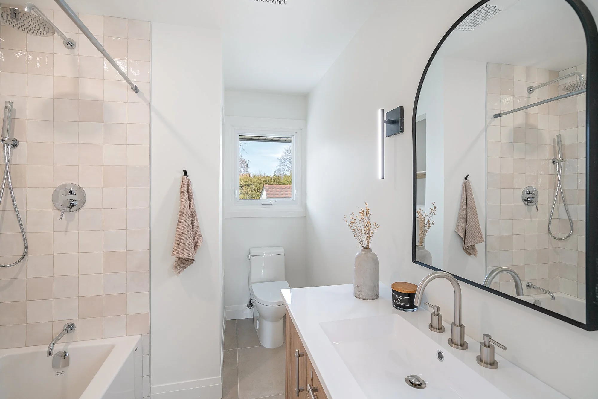Modern bathroom with white walls, a window, a bathtub with a shower, a toilet, a wooden vanity with a white countertop, a large mirror, and decorative vases.