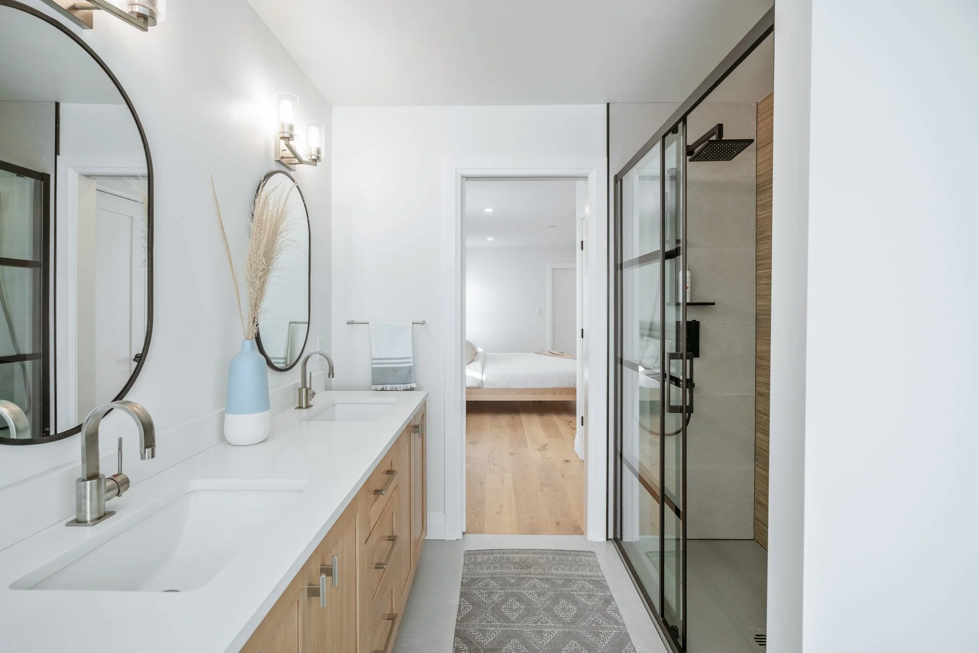 Modern bathroom with dual sinks, a shower with glass doors, and a view into a bedroom with a bed and hardwood floors.