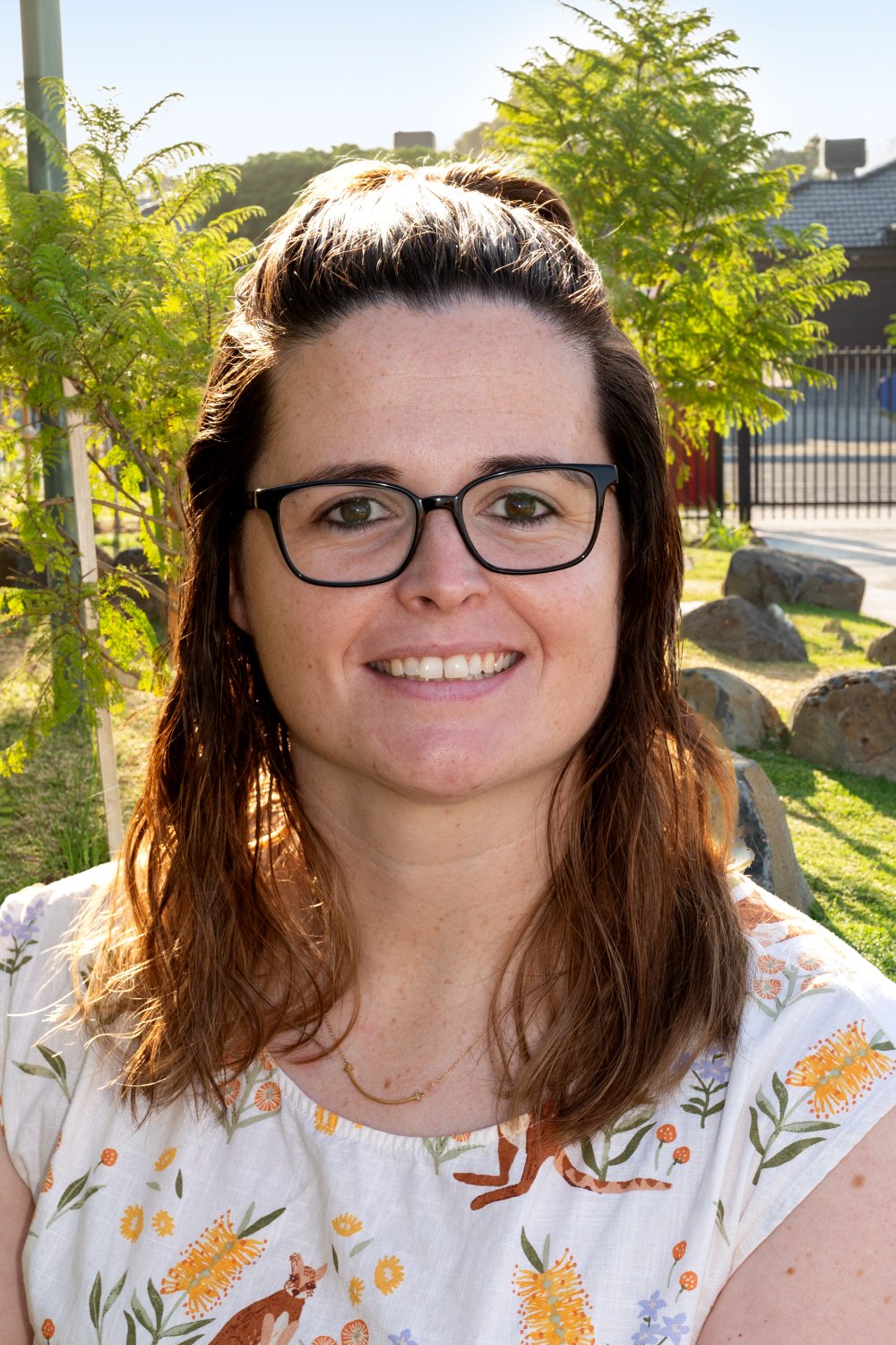 Smiling woman with brown hair, glasses, and a floral-patterned white top, outdoors near greenery and rocks, in bright sunlight.