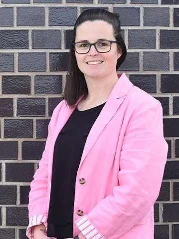 A woman with dark hair, wearing glasses, a pink blazer, and a black top, standing in front of a black brick wall and smiling.