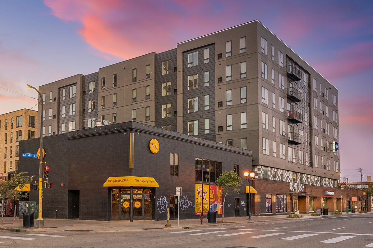 A mixed-use building with a restaurant on the ground floor and apartment units above, located at the corner of an intersection during sunset, with colorful sky and streetlights.