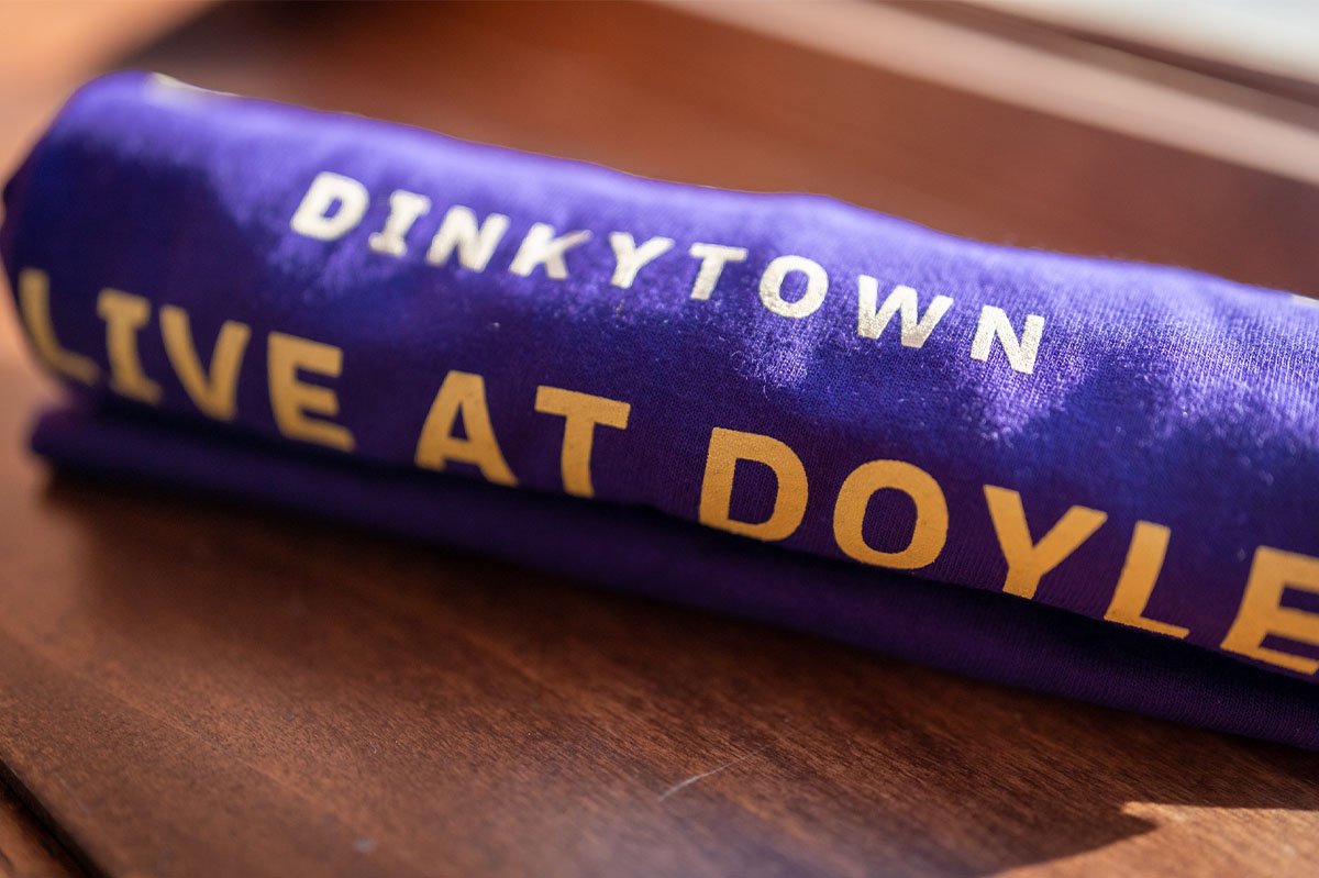Purple T-shirt with the phrase 'DINKYTOWN' and part of the phrase 'LIVE AT DOYLE' printed on it.