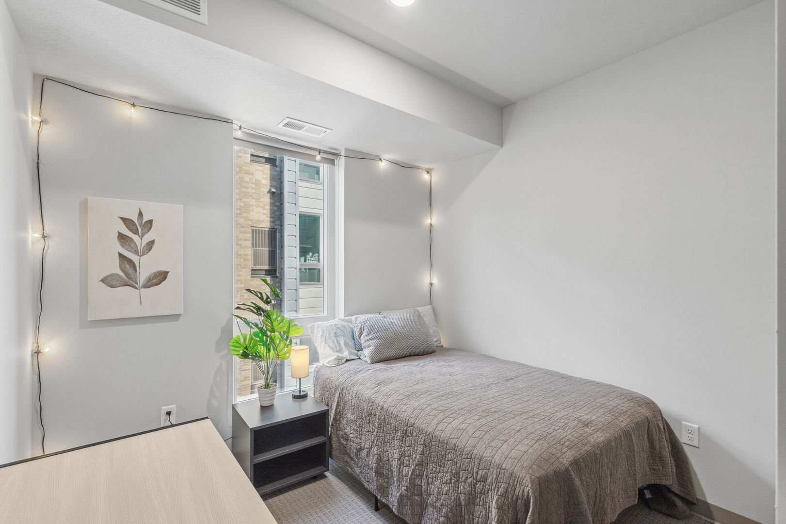 Bedroom with a bed, nightstand with a lamp and plant, window with outside view, and string lights on white walls.
