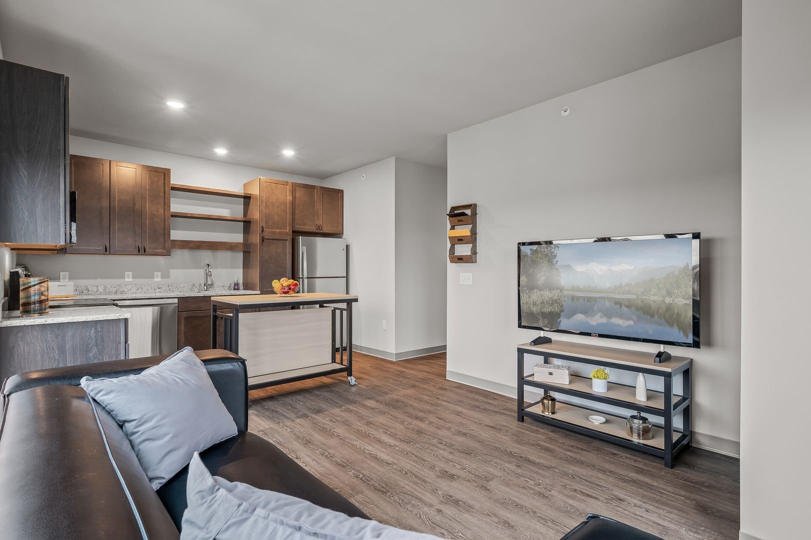 Living room and kitchen area with gray walls, dark wood flooring, a black leather sofa with pillows, a flat-screen TV on a black and wood stand, and kitchen cabinets with a small kitchen island.