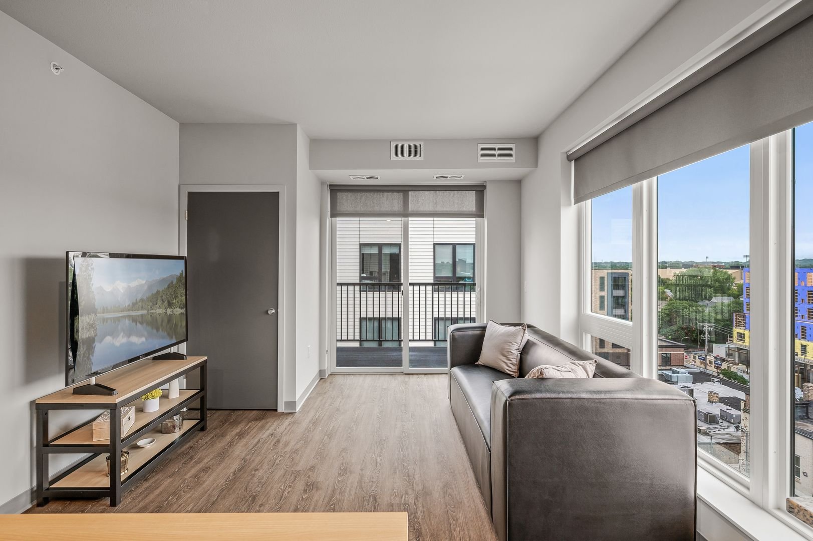 Living room with a gray sofa, large windows with roller shades, wooden flooring, TV on a stand, and a balcony visible through sliding glass doors.