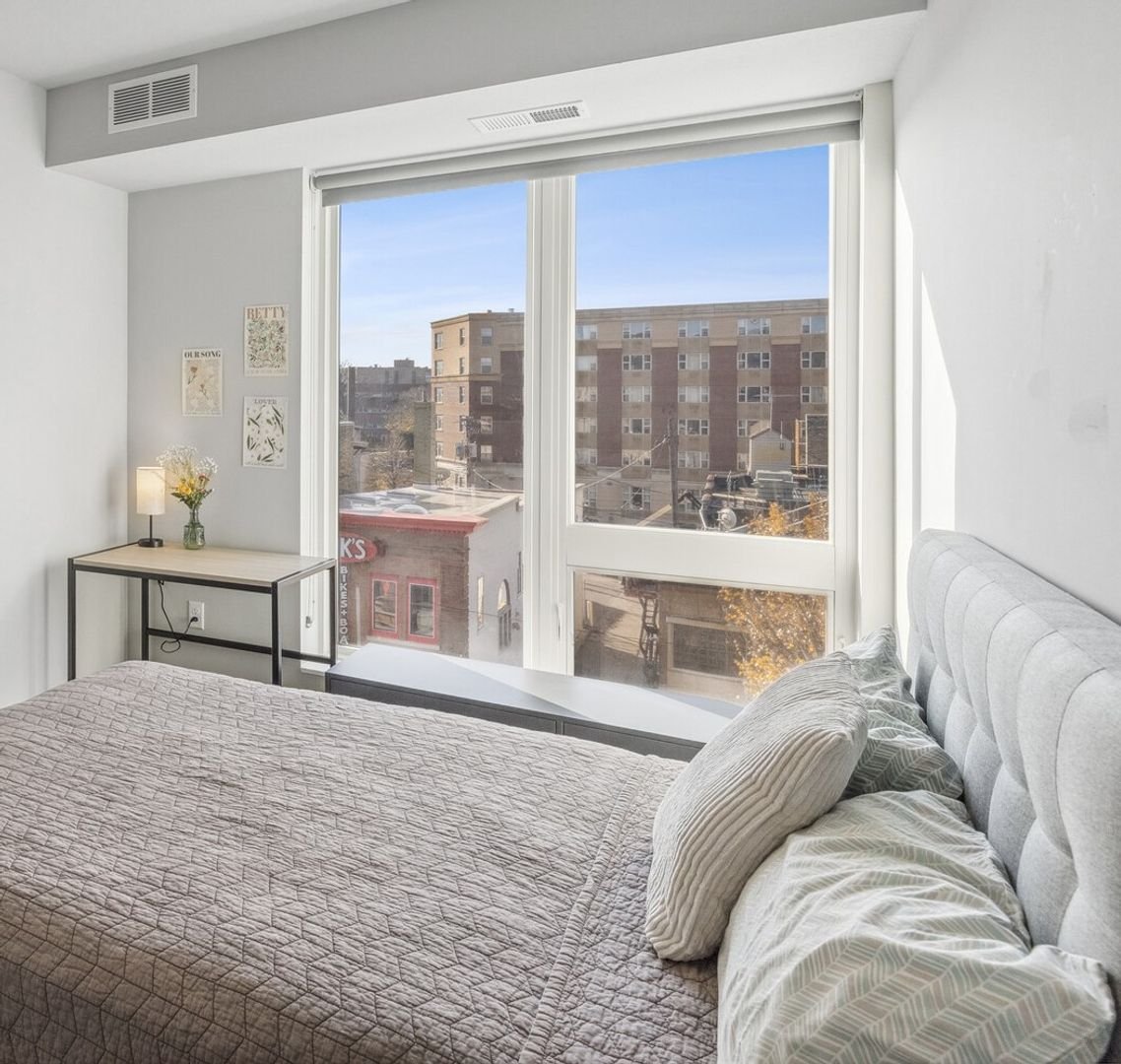 A bedroom with a large window showing a city view, a bed with a quilted beige bedspread, pillows, a small black metal desk with a lamp and flower vase, and framed artwork on the wall.