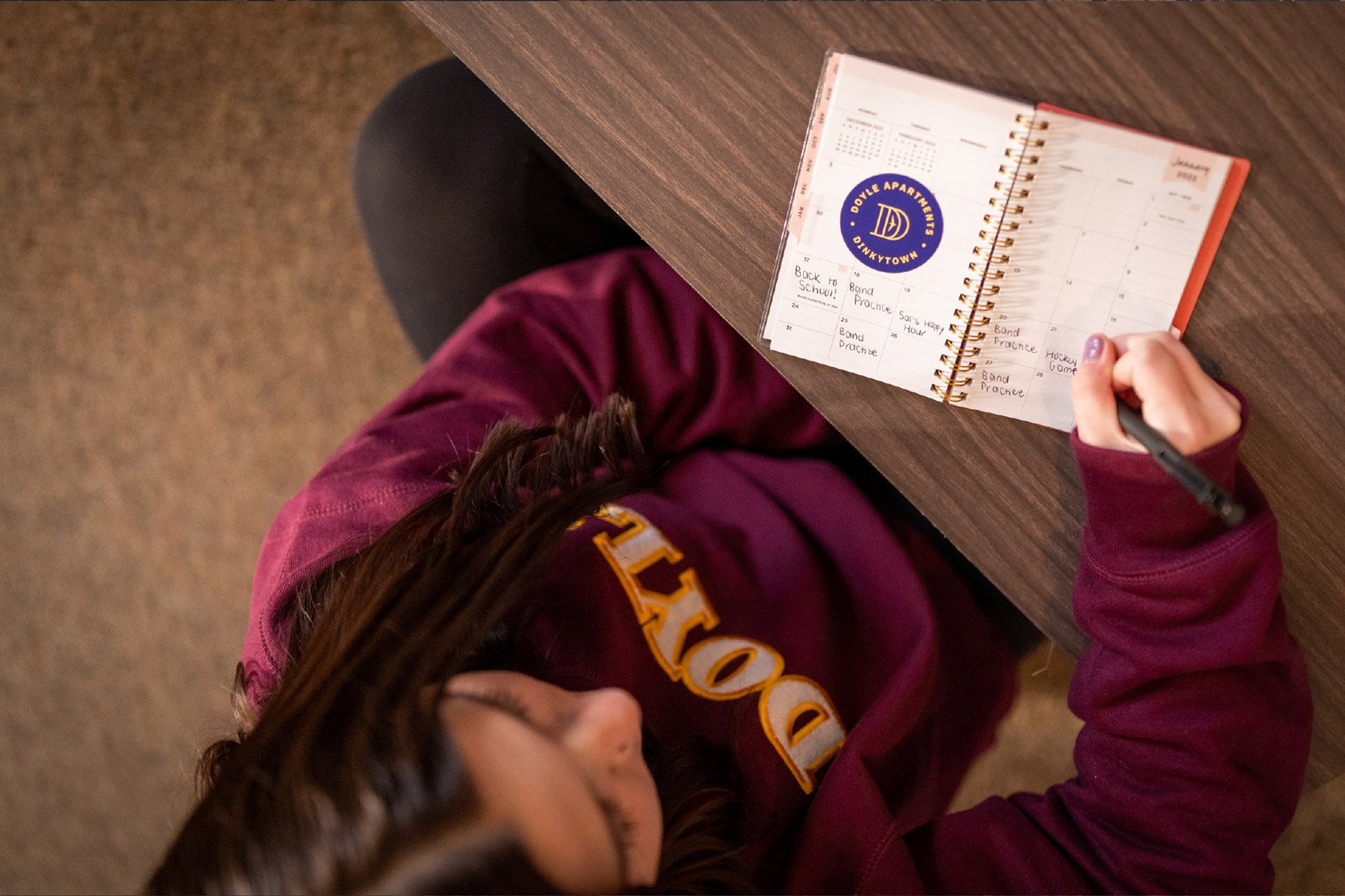 Overhead view of a person writing in a planner at a desk, with a band practice schedule, wearing a maroon hoodie with yellow lettering.