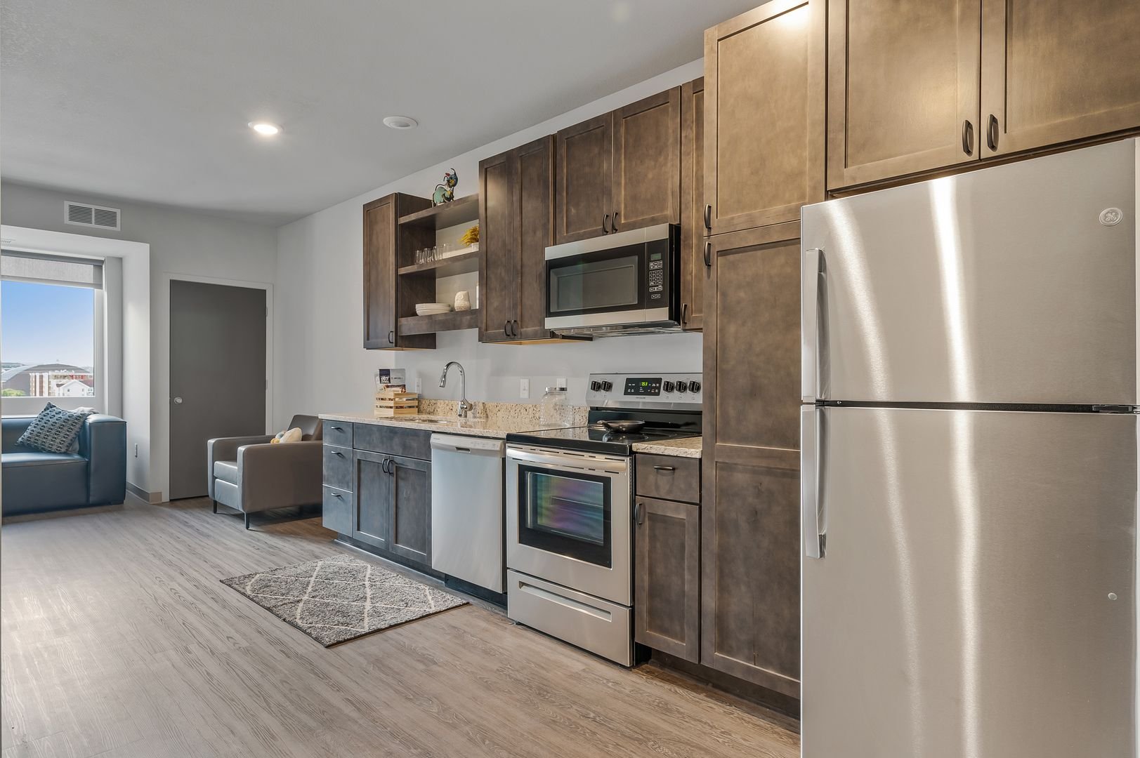 Kitchen with brown cabinets, stainless steel refrigerator, microwave, oven, and a white dishwasher. Light wood flooring and a window with a view of neighboring buildings.