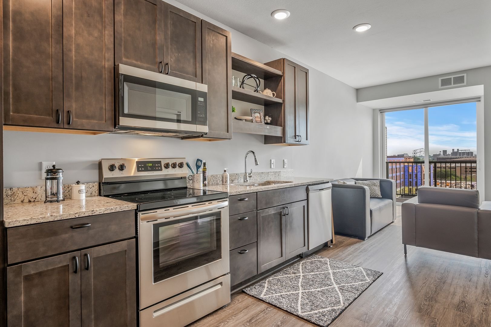 Modern apartment kitchen with dark wood cabinets, stainless steel appliances, granite countertops, and a living area with gray furniture and a large window showing a Minneapolis view.