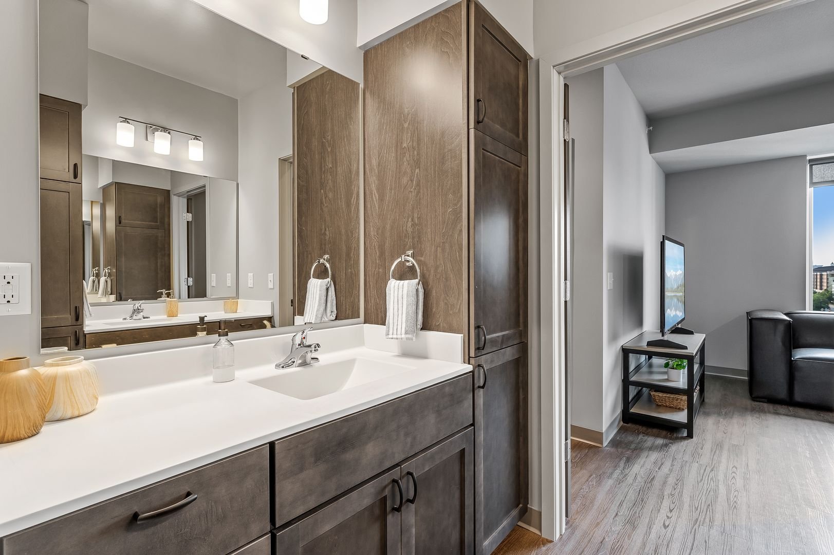 Bathroom vanity with mirror, towels, and soap dispenser next to a doorway leading to a living room with a TV, black sofa, and window.