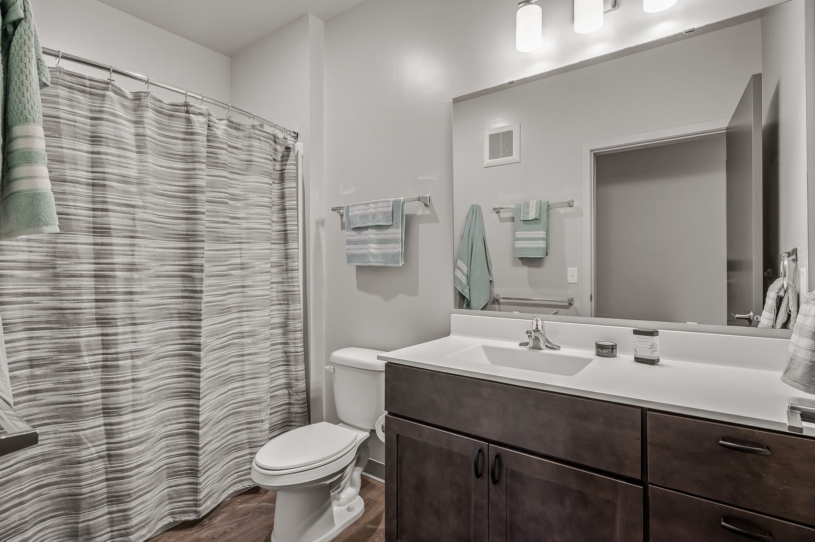 Bathroom with a shower, a toilet, and a vanity with a large mirror, dark wood cabinet, potted soap, and towels on the wall, lit by three ceiling lights.