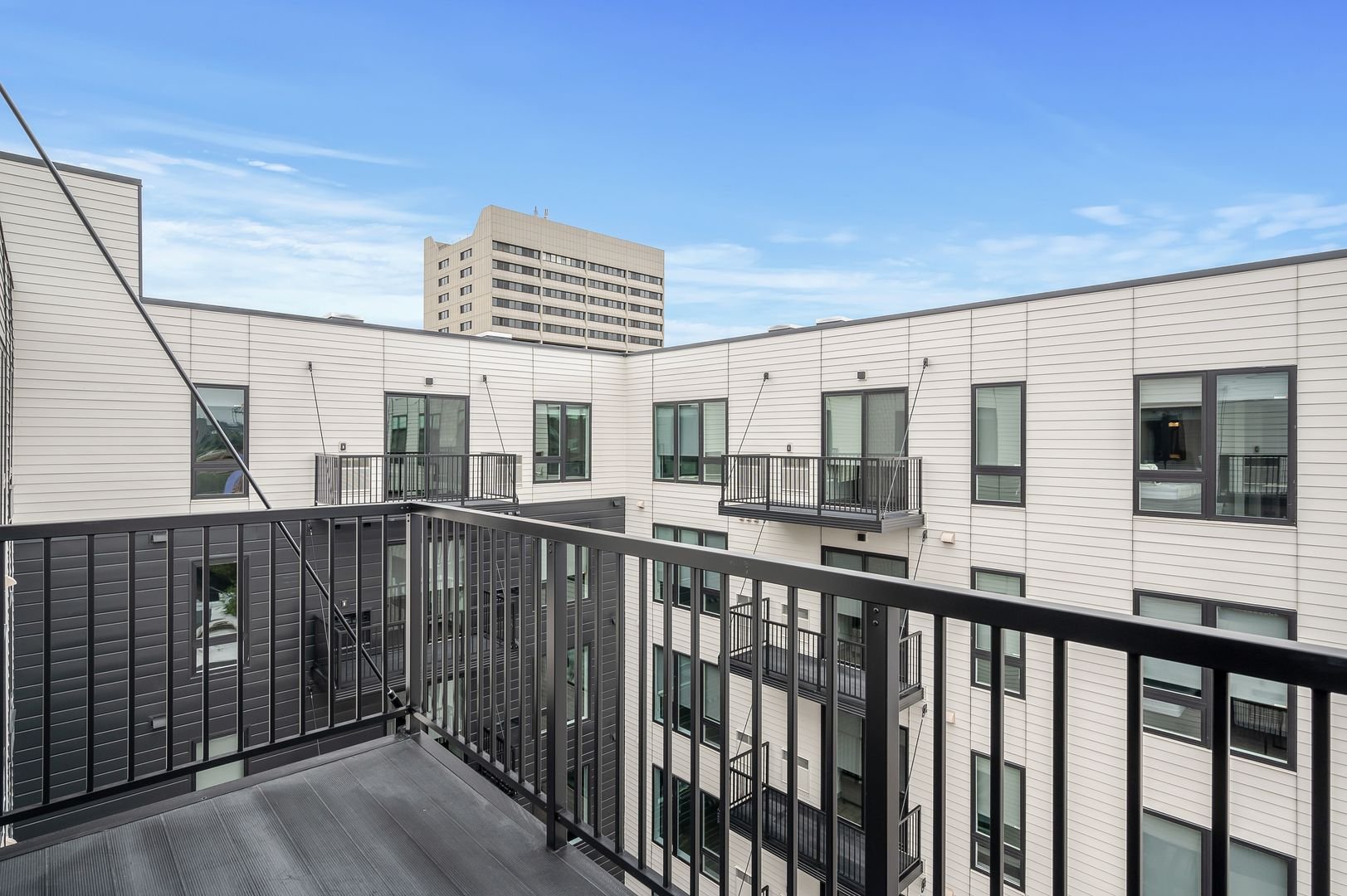 View from a balcony of a modern apartment building with multiple small balconies, large windows, and a taller building in the background under a blue sky.