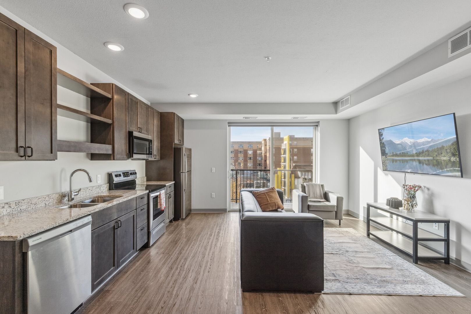 Open-concept living room and kitchen in an apartment with wooden flooring, a balcony with a view of nearby buildings, a flat-screen TV, sofa, armchair, and modern kitchen appliances.