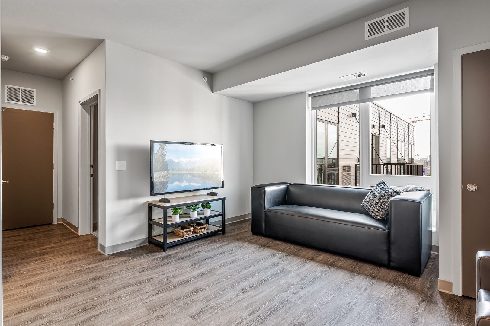 Living room with wooden floor, black sofa, TV, and large window showing an outdoor balcony with railing and building exterior.
