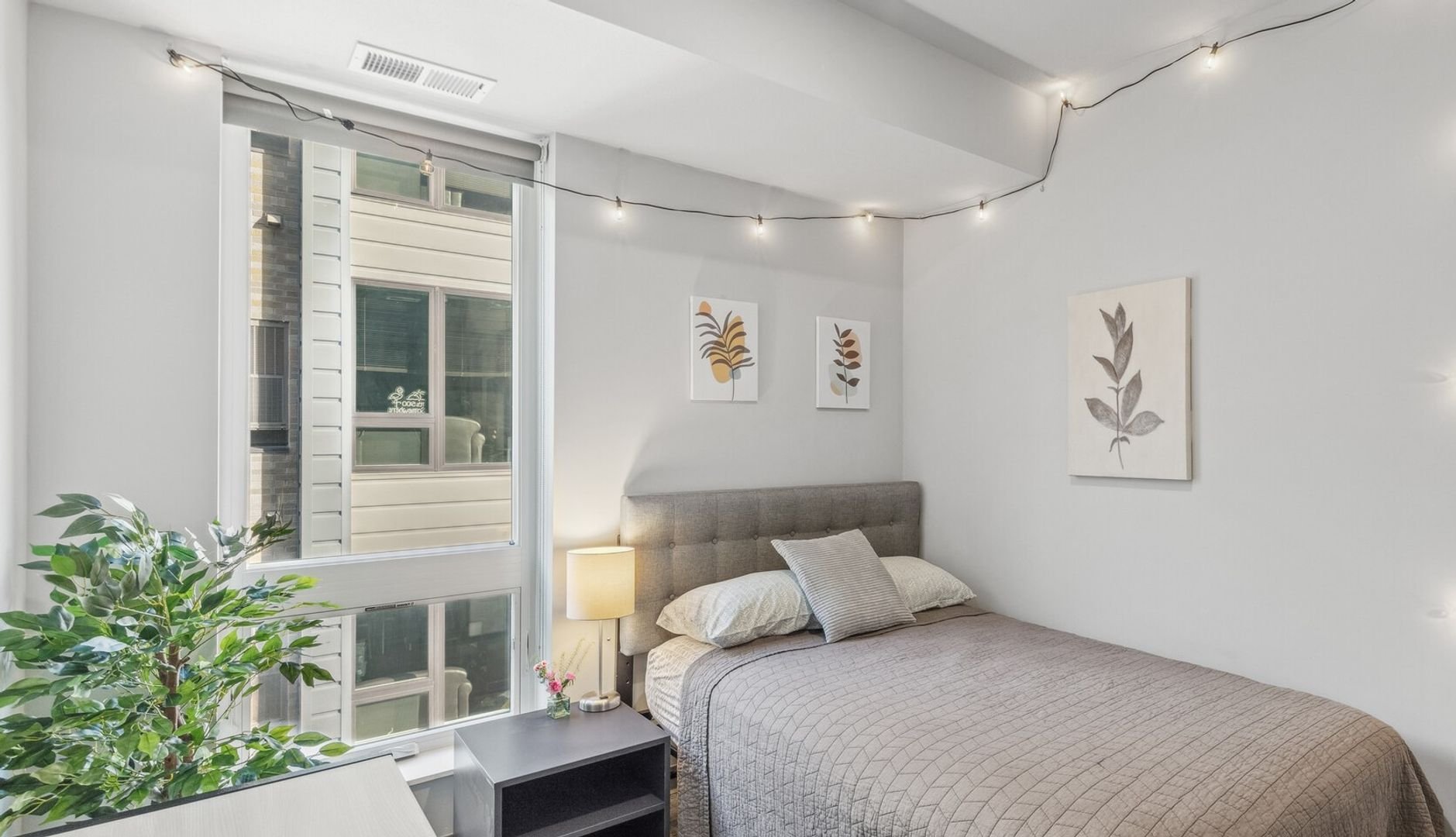 A cozy bedroom with a large window, a bed with gray headboard, beige bedding, and decorative pillows, a nightstand with a lamp.