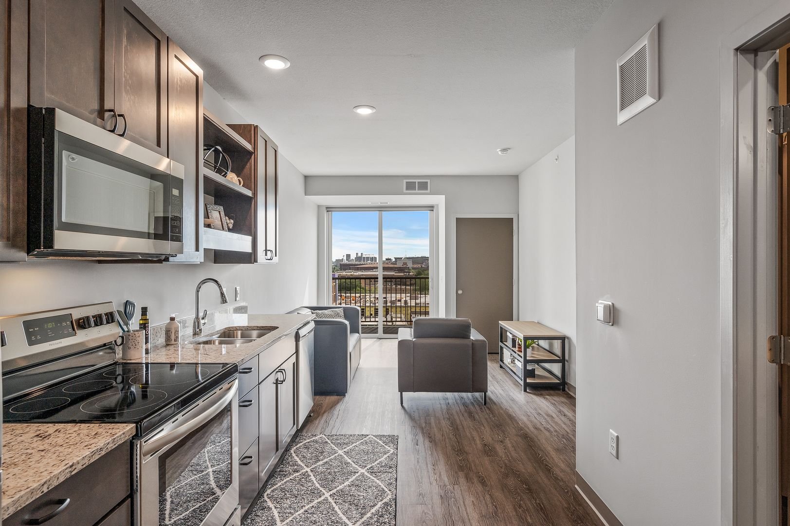 Modern kitchen and living room area with wood flooring, gray and black furniture, and a sliding glass door leading to a balcony, with Minneapolis skyline in the distance.