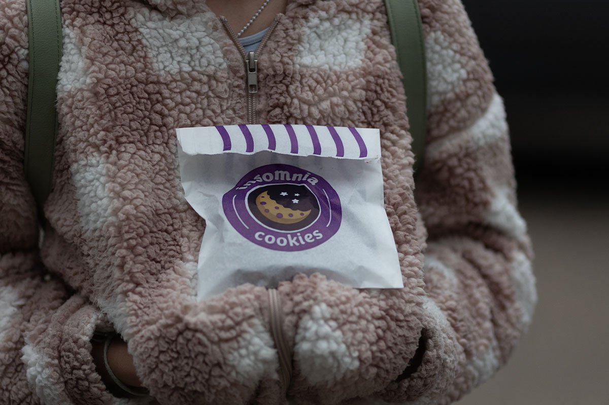 Person in a fluffy pink and white zip-up jacket holding a white paper bag of cookies with a purple circular logo that says "Insomnia Cookies".