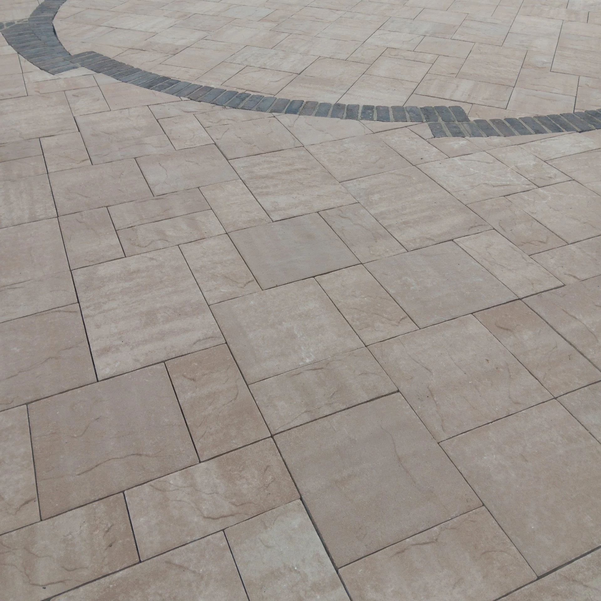 A paved surface with large beige rectangular tiles and a curved border made of dark gray bricks.