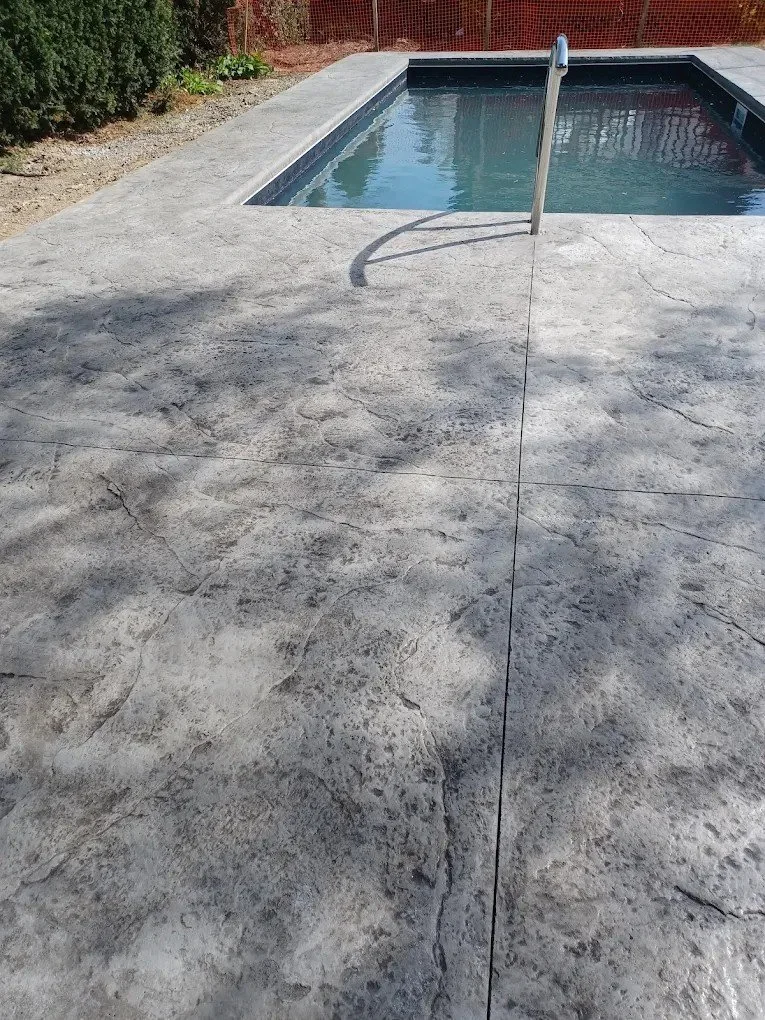 A small rectangular swimming pool with a metal handrail, surrounded by a textured gray concrete deck, with bushes and a red fence in the background.