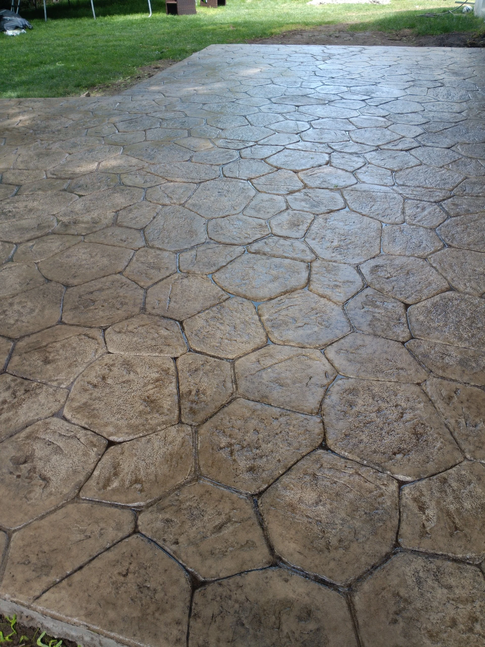 A newly paved concrete patio area with a pattern of irregular, hexagonal shapes in a backyard with green grass.