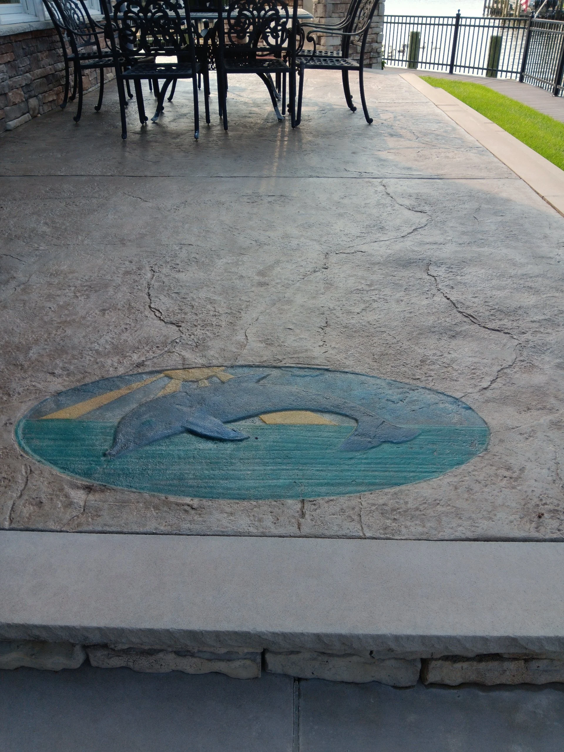 Painted dolphin on concrete floor under outdoor patio area with black metal table and chairs, and a railing overlooking water.