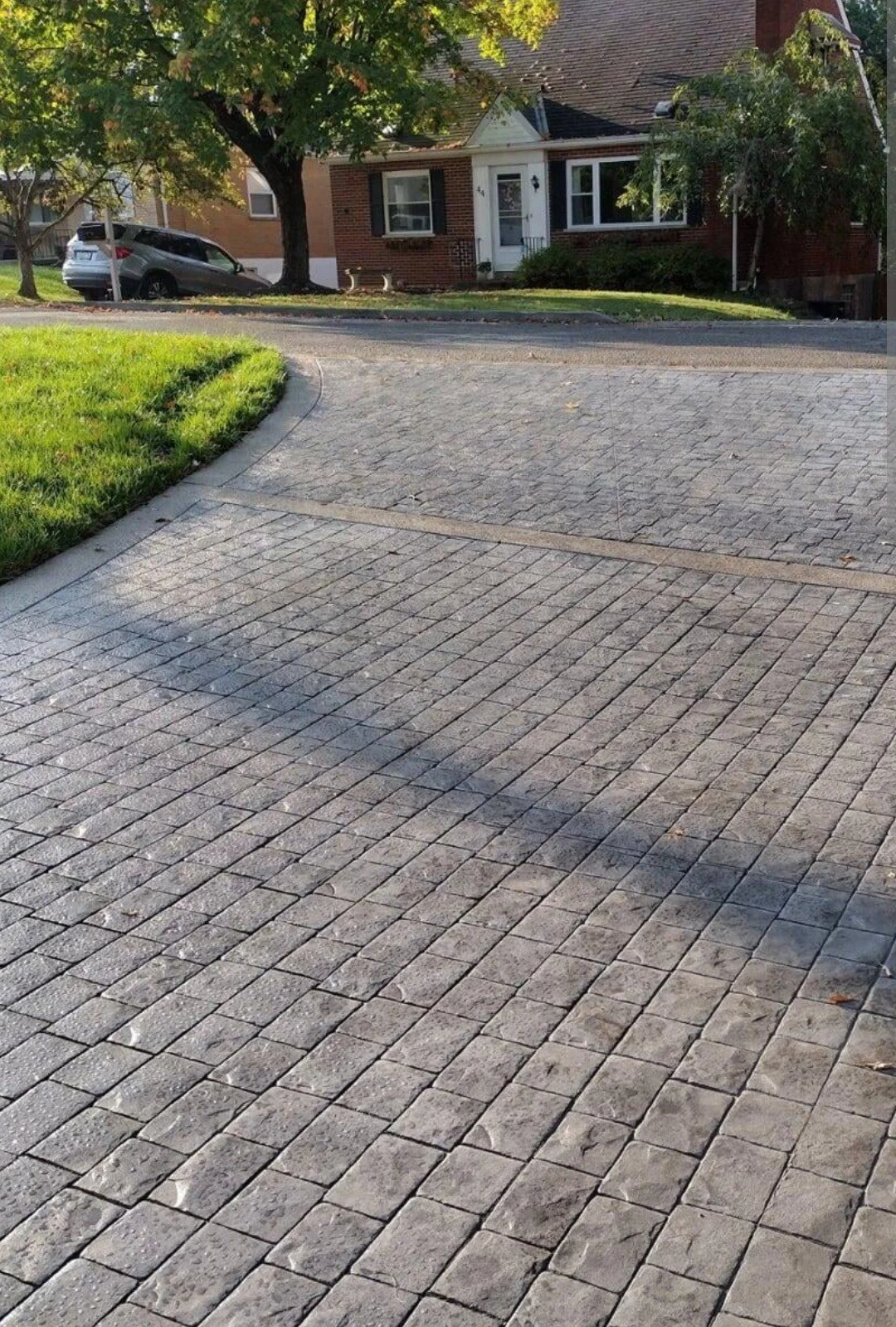 Residential street with brick house, parked cars, large trees, and a stone driveway.