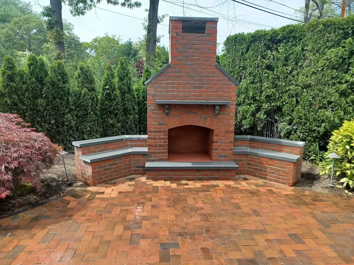 Red brick outdoor fireplace with a chimney, built-in benches on either side, and a paved brick patio in a landscaped backyard with trees and shrubs.