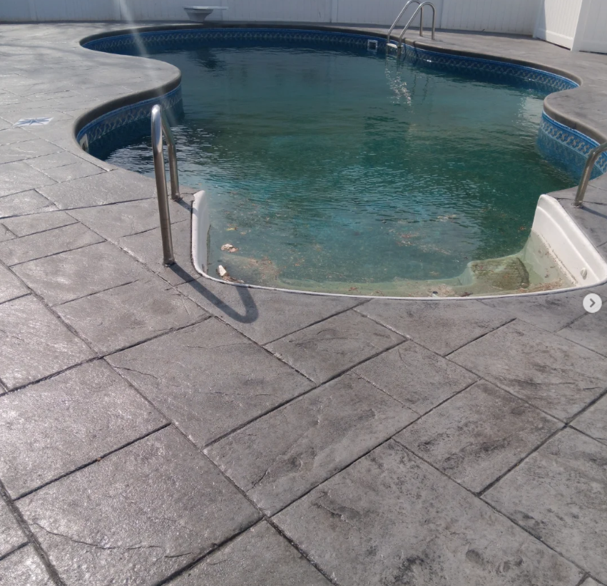 An outdoor swimming pool with dirty, algae-covered water and a concrete patio surrounding it. The pool has metal handrails and a curved entry steps area.