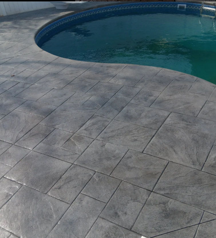 A backyard with a concrete pool deck surrounding a swimming pool filled with water.