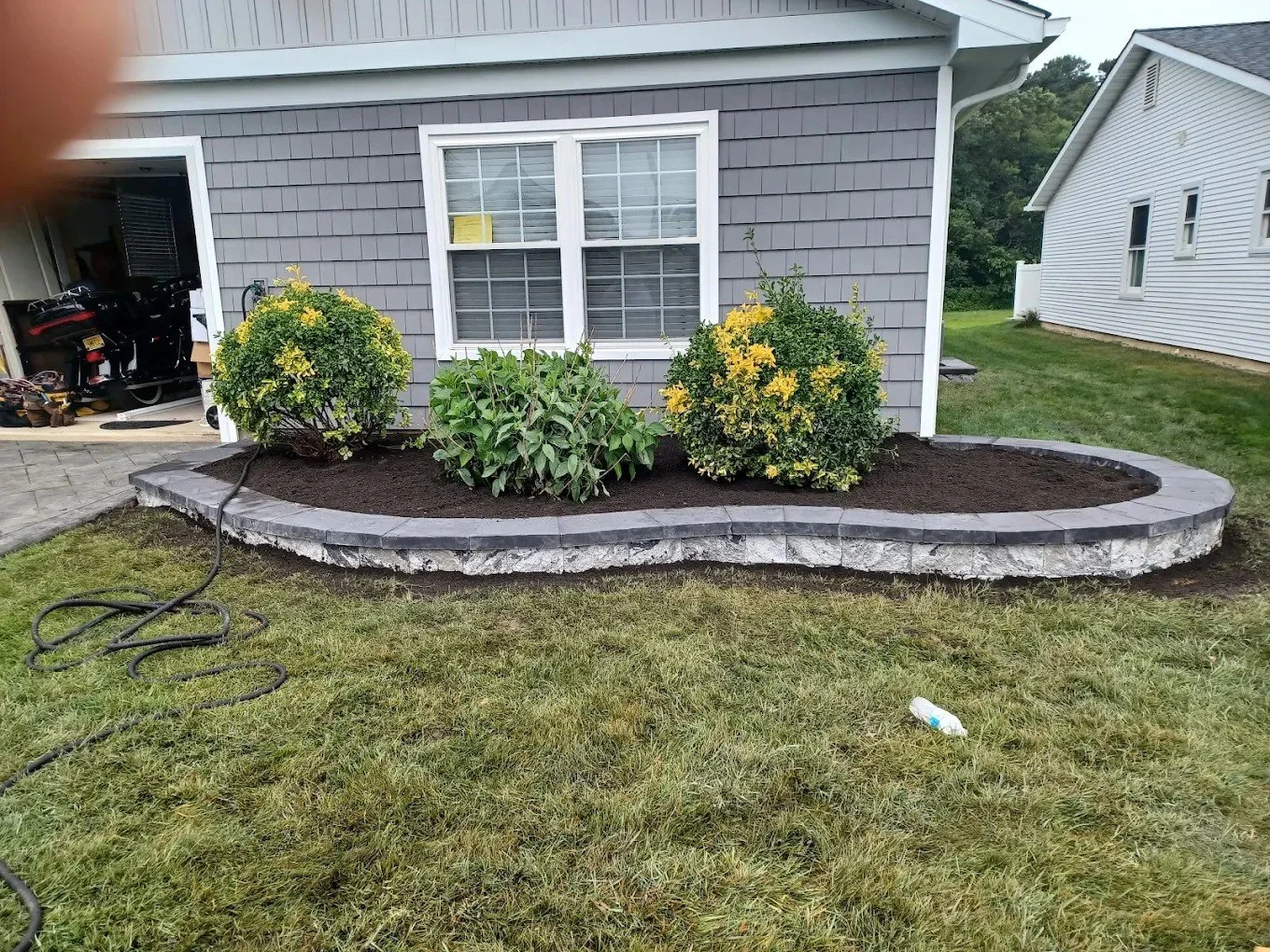 A garden bed with dark soil, bordered by stone bricks, with three bushes planted in front of a house with gray siding. A window with white trim is visible on the house, and there is a hose lying on the grass.