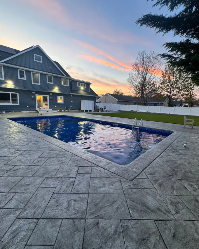 Residential backyard with a blue swimming pool, gray stone patio, white fence, a blue house with multiple windows, trees, and a sunset sky with pink and orange clouds.