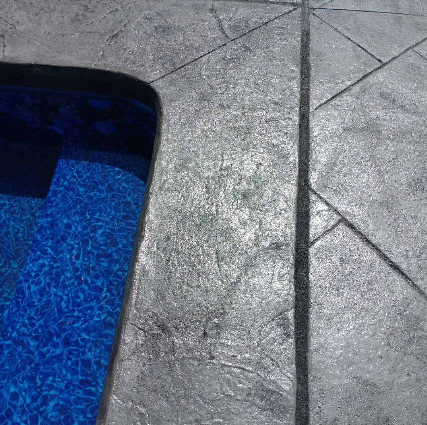 Corner of a swimming pool with blue water next to textured gray concrete pool deck.