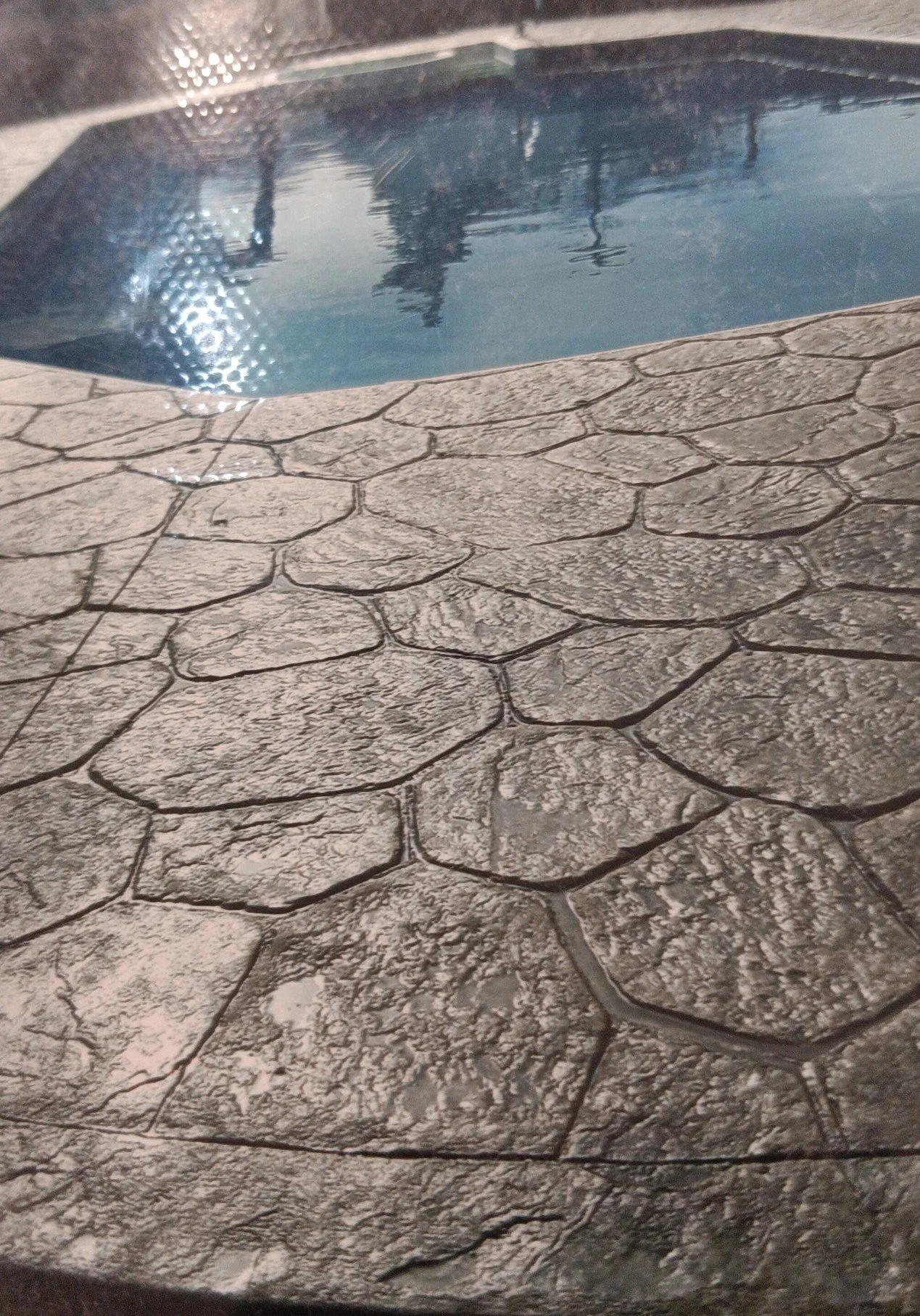 Close-up of textured stone pool deck with a small pool of water reflecting surroundings.