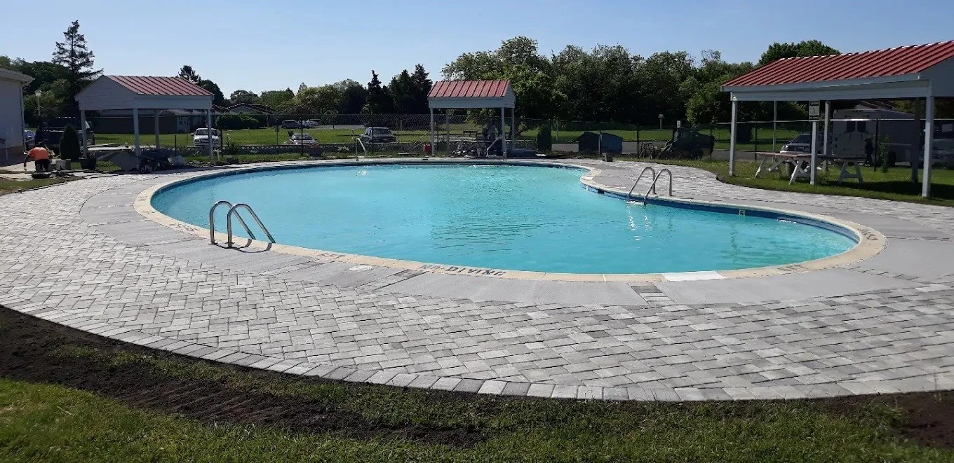 Empty outdoor swimming pool with paved surround, surrounded by grassy areas, small shelter structures, and parked cars in the background.
