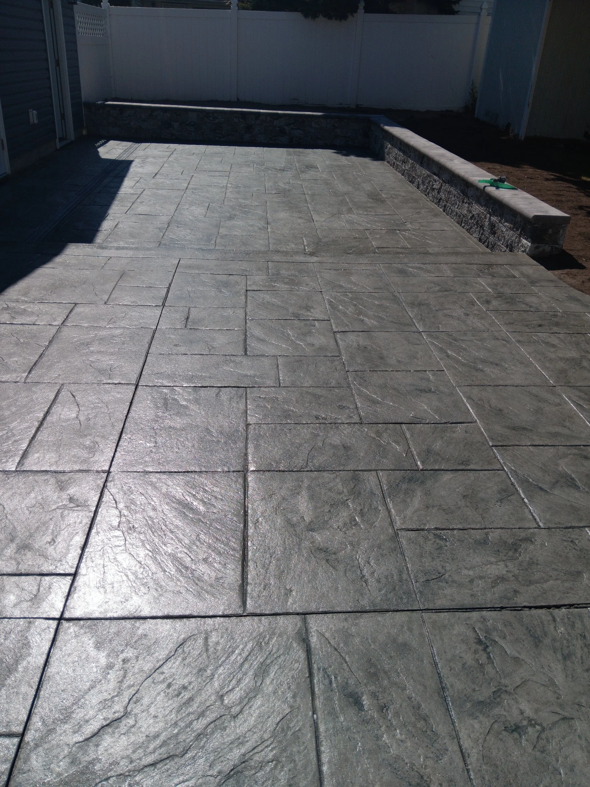 Backyard patio with gray concrete stamped concrete flooring, a white privacy fence, and a low stone retaining wall with a raised garden bed.