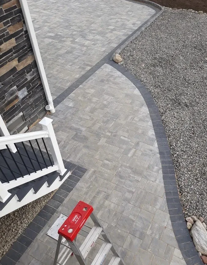 Concrete and stone pathway with a black and gray border, next to a brick wall, with a white staircase and a red step stool outside.