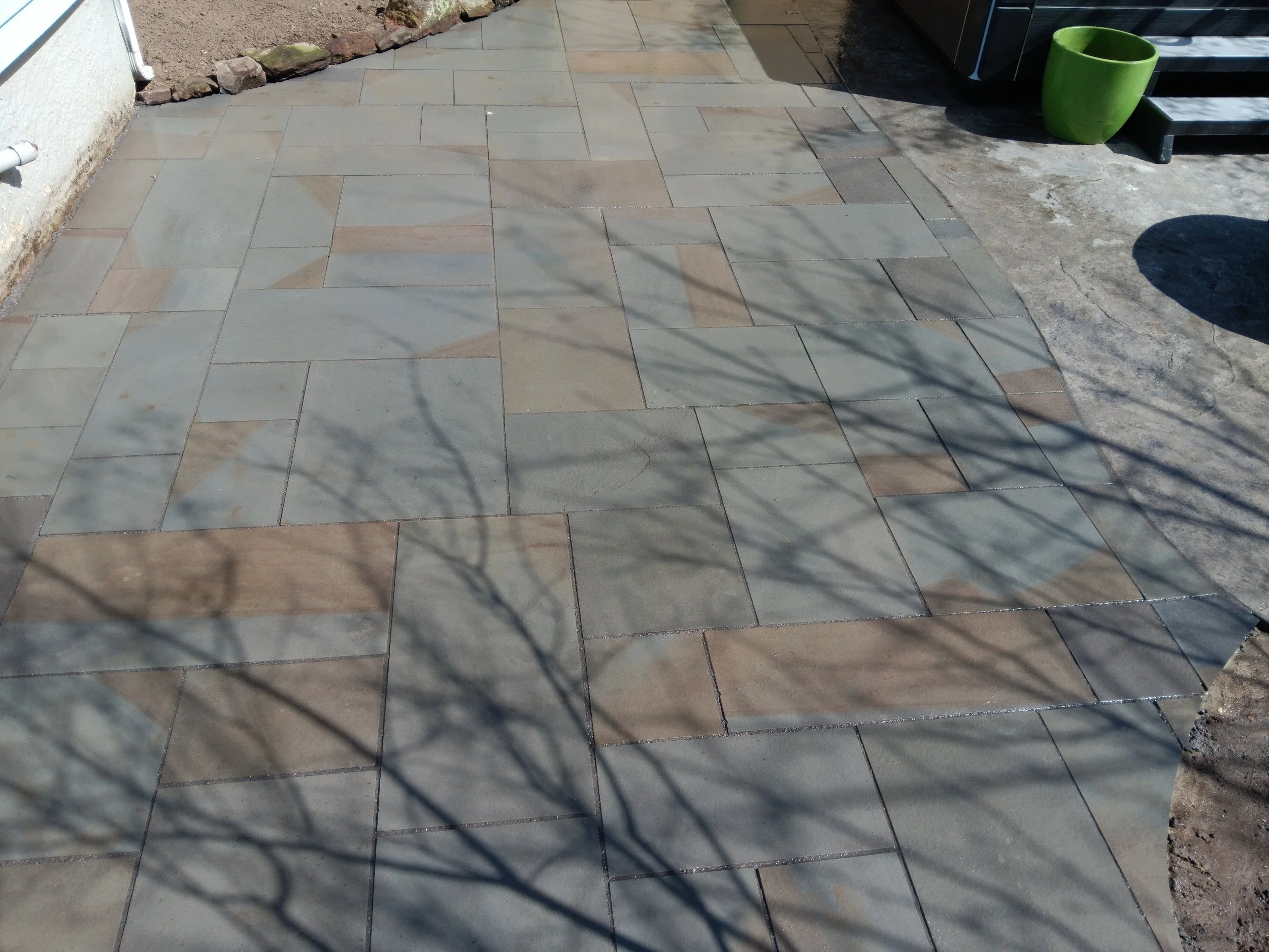 Newly installed stone patio with multicolored rectangular tiles, next to a white house wall and a green flowerpot, with shadows of trees cast across the surface.