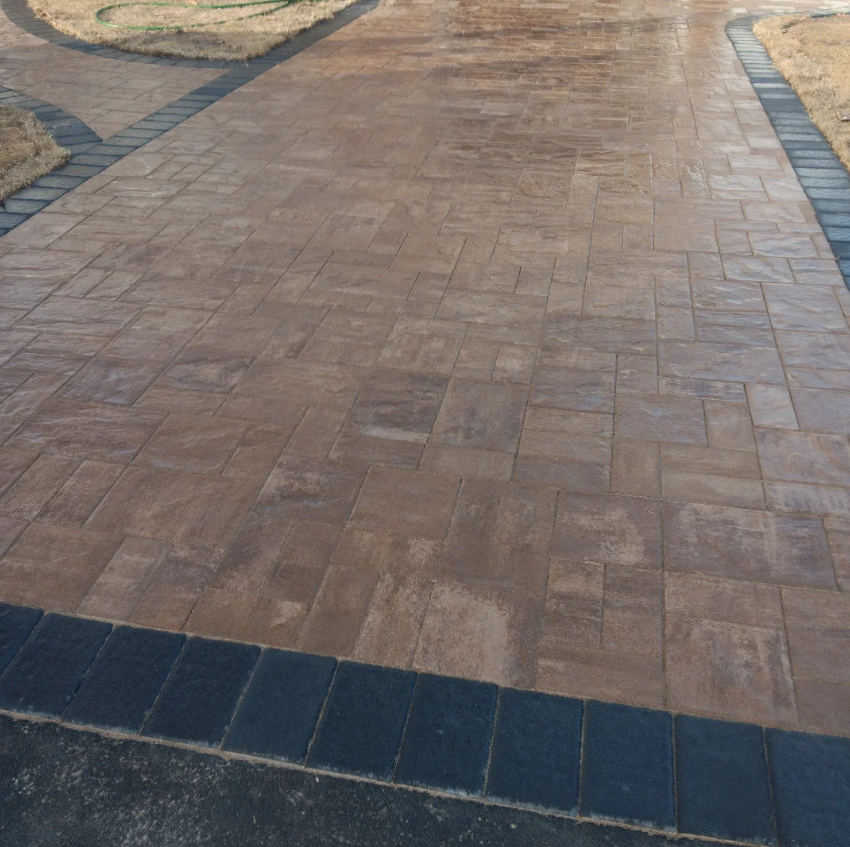 A paved walkway made of brown concrete tiles with a border of dark gray bricks, surrounded by patches of grass and dirt.