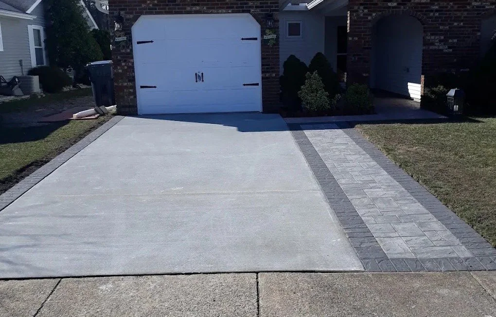 Concrete driveway with a brick border leading to a white garage door surrounded by brick walls, with a sidewalk and some grass on the sides.