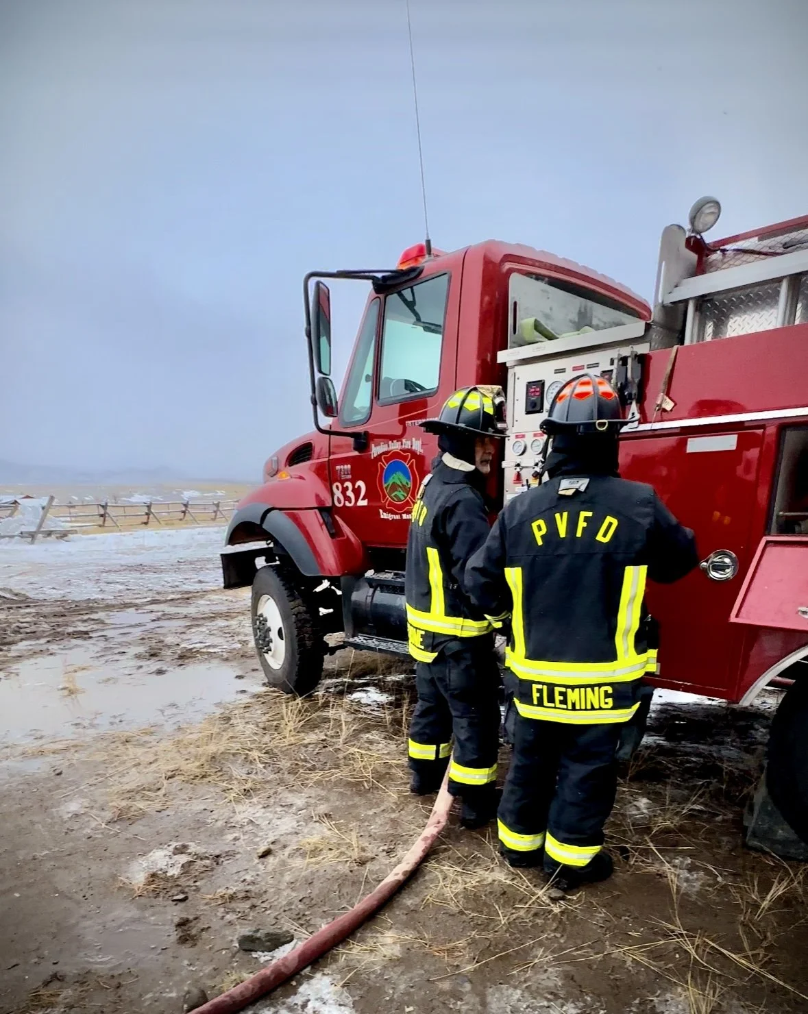 Firefighters in black gear and helmets stand next to a red fire truck at the beach during overcast weather.