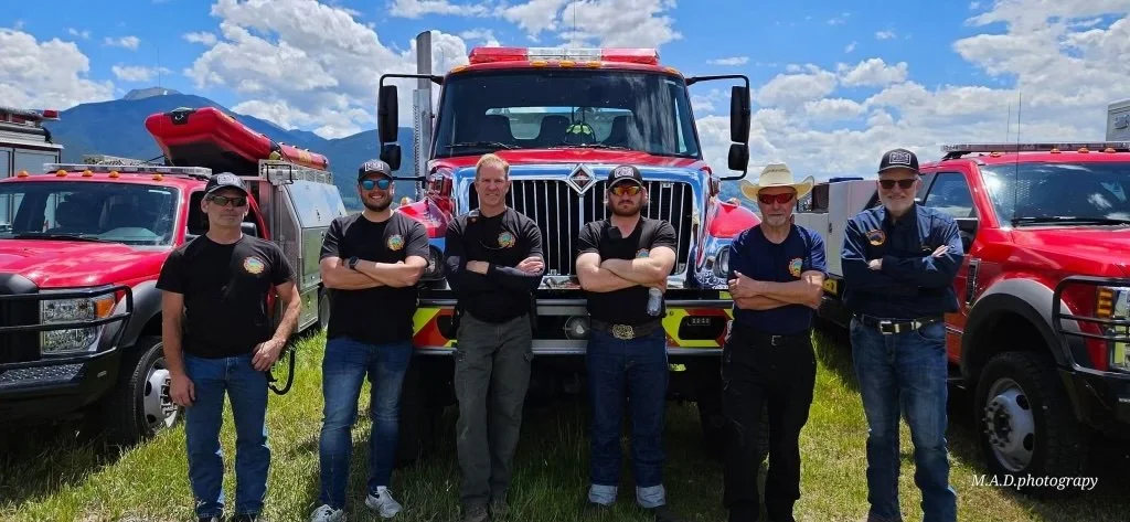 Group of six firefighters standing in front of fire trucks on grassy field under cloudy sky