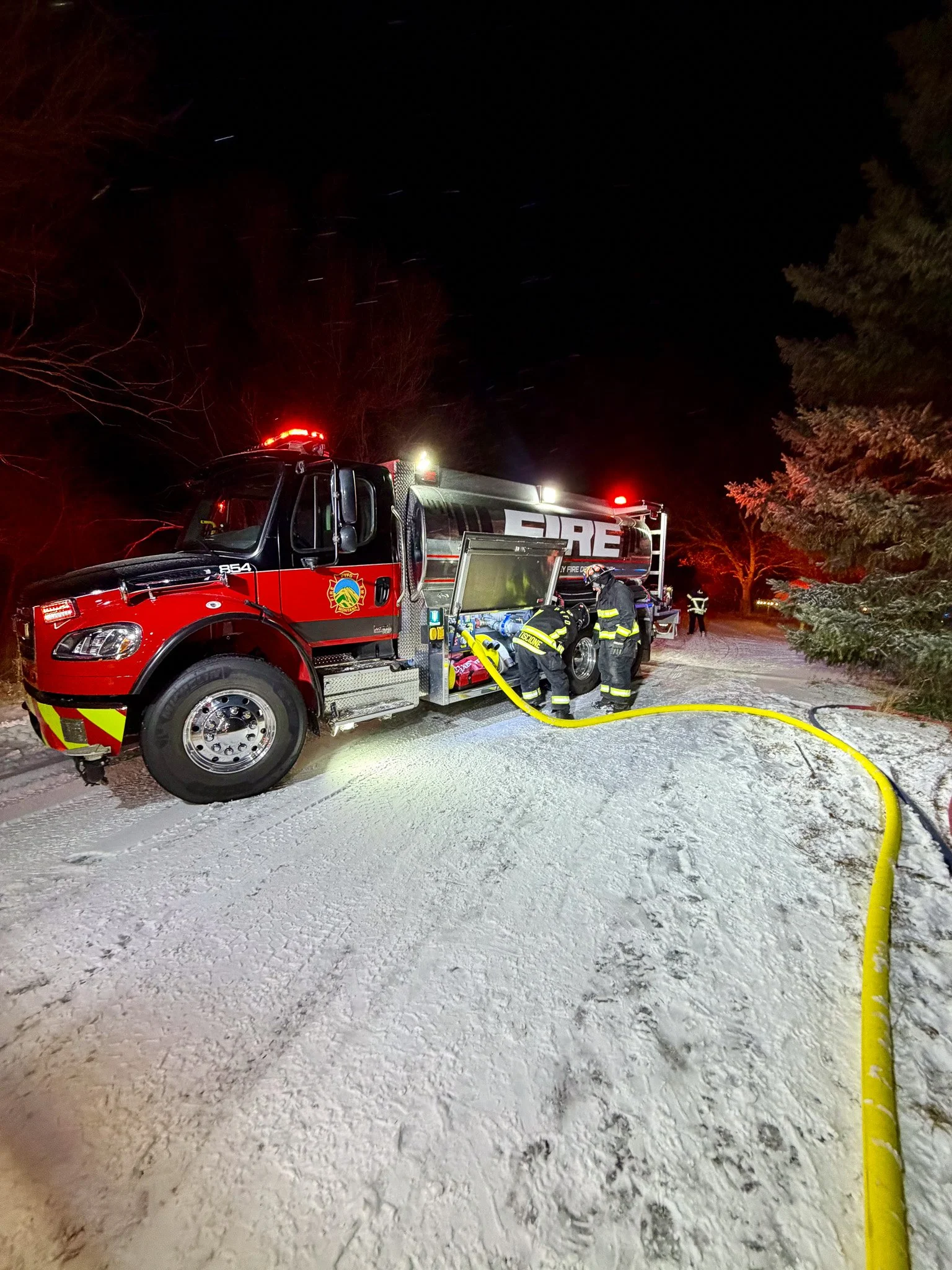 Firefighters fueling a fire truck at night on a snowy ground, with trees and emergency lights in the background.