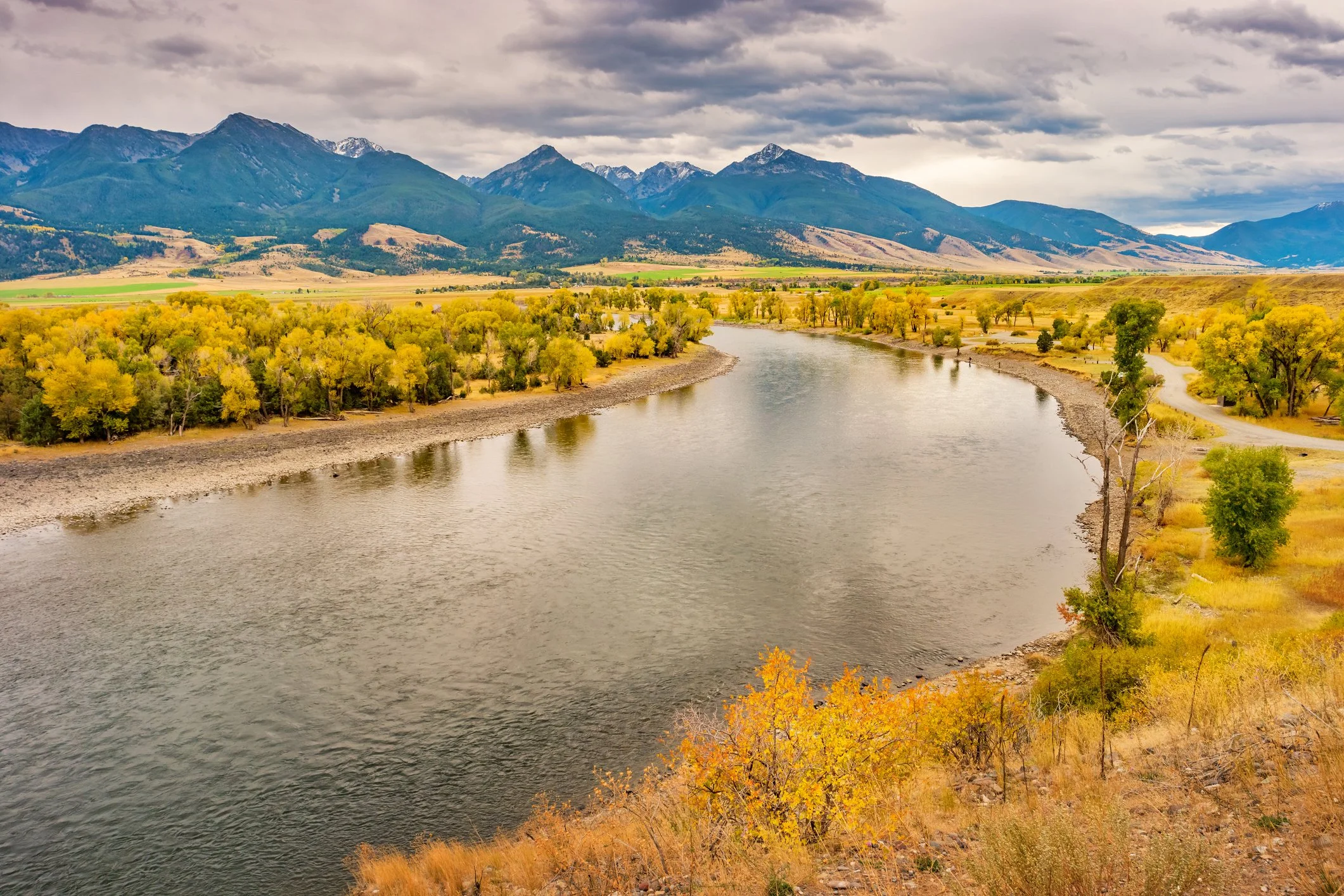 A river flowing through a valley with yellow and green trees on its banks, and mountains with snow-capped peaks in the background under a cloudy sky.
