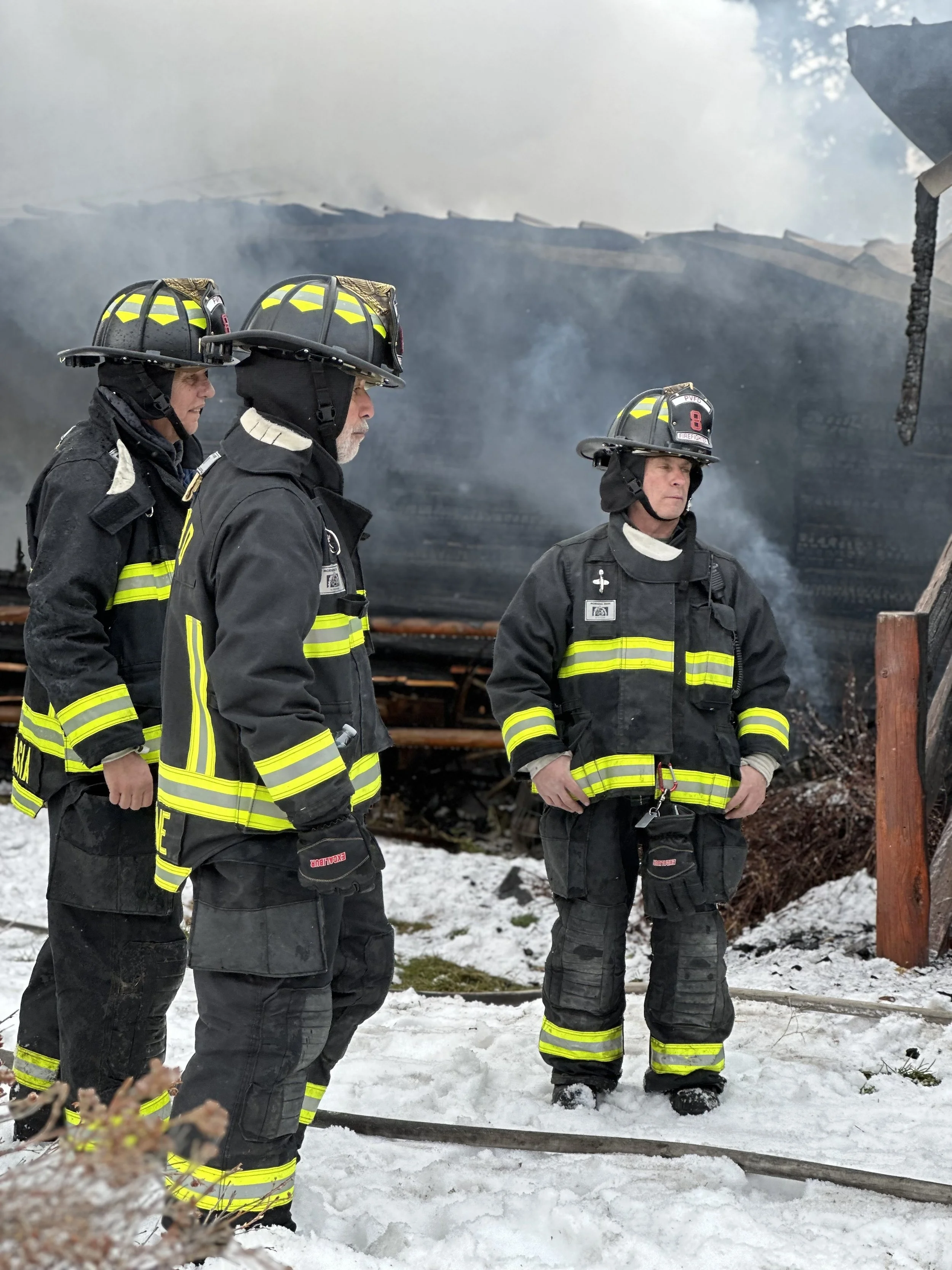 Four firefighters in black turnout gear with reflective yellow stripes, wearing helmets, standing on snow-covered ground in front of a smoldering burnt building.
