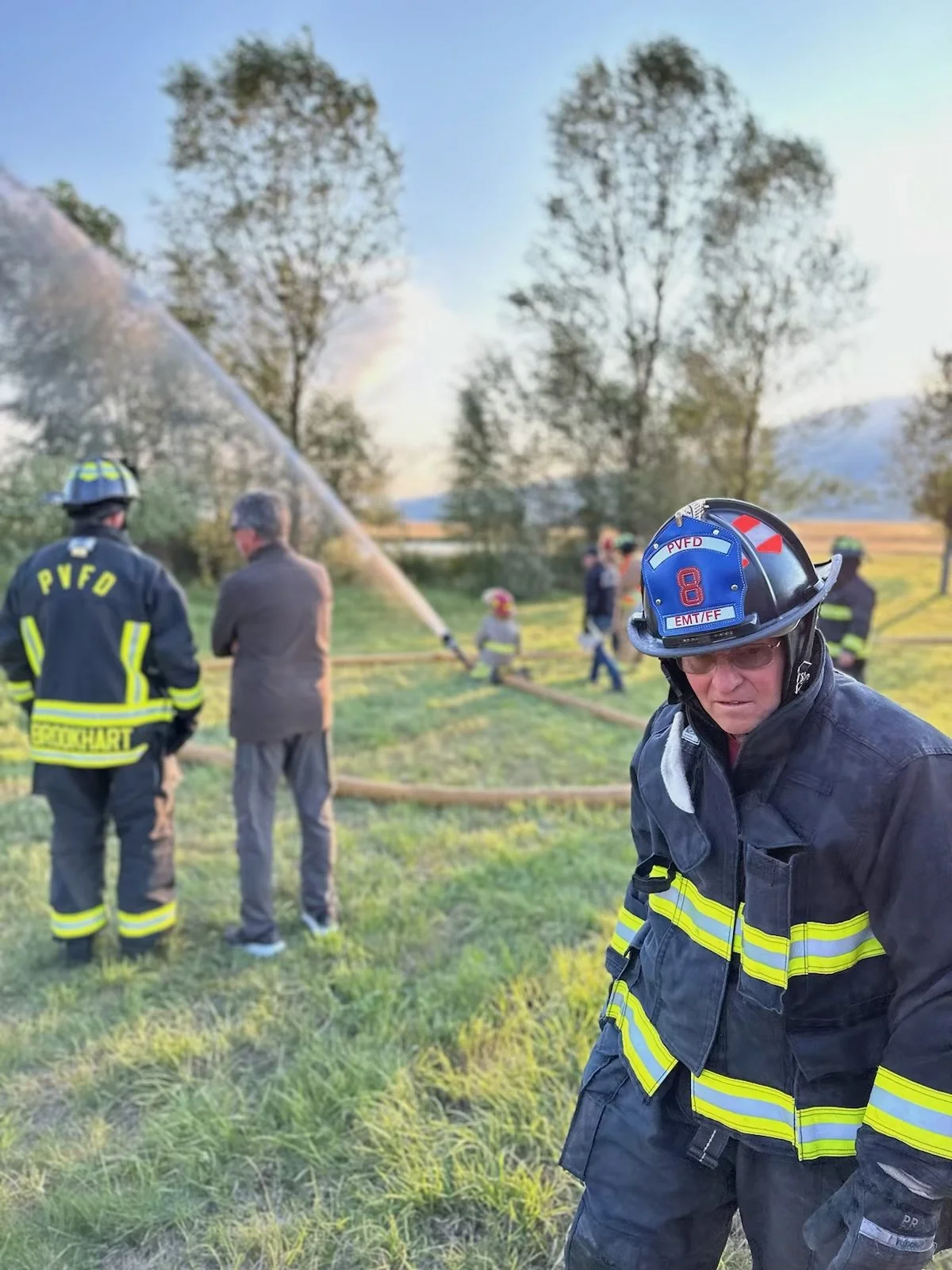 Firefighters and officials at an outdoor scene with a firehose spraying water, trees in the background, and a firefighter in the foreground wearing a helmet and uniform.