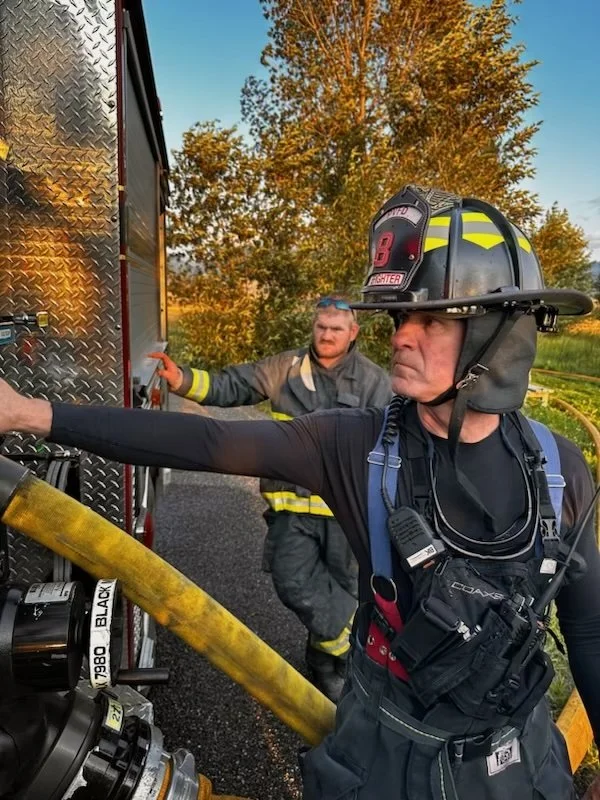 Firefighters preparing to fight a fire, one firefighter is holding a fire hose with a yellow nozzle and pointing it forward, wearing a helmet and gear, with another firefighter in the background.