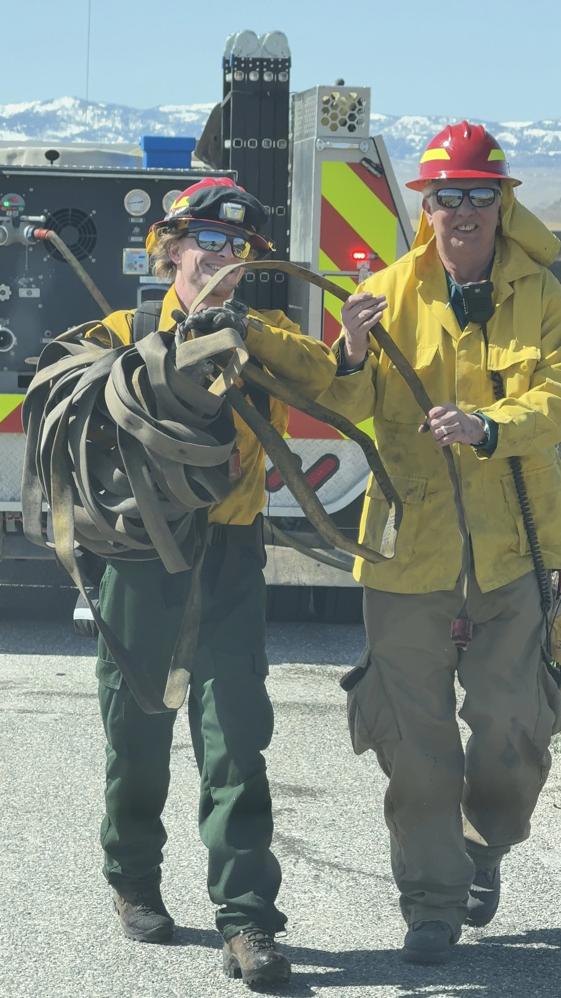 Two firefighters in yellow gear and helmets, one holding a tangled hose, standing in front of a fire truck with mountains in the background.